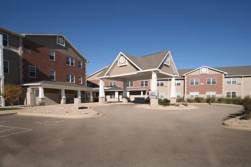 Exterior view of Oak Park Place Dubuque, showing a large three-story building with a covered entrance supported by white pillars. The building features a combination of red brick and beige siding under a clear blue sky.
