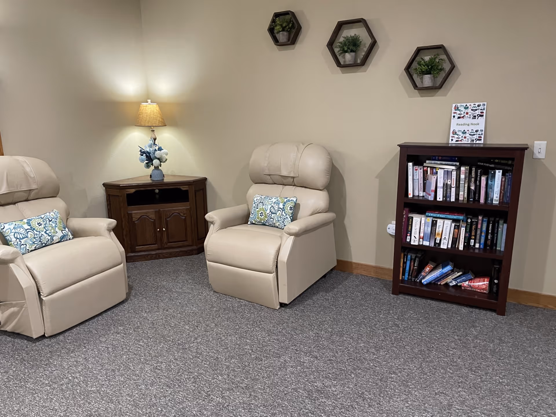 Two beige recliners with patterned pillows in a small reading area featuring a lamp on a corner table, wall shelves, and a bookshelf.