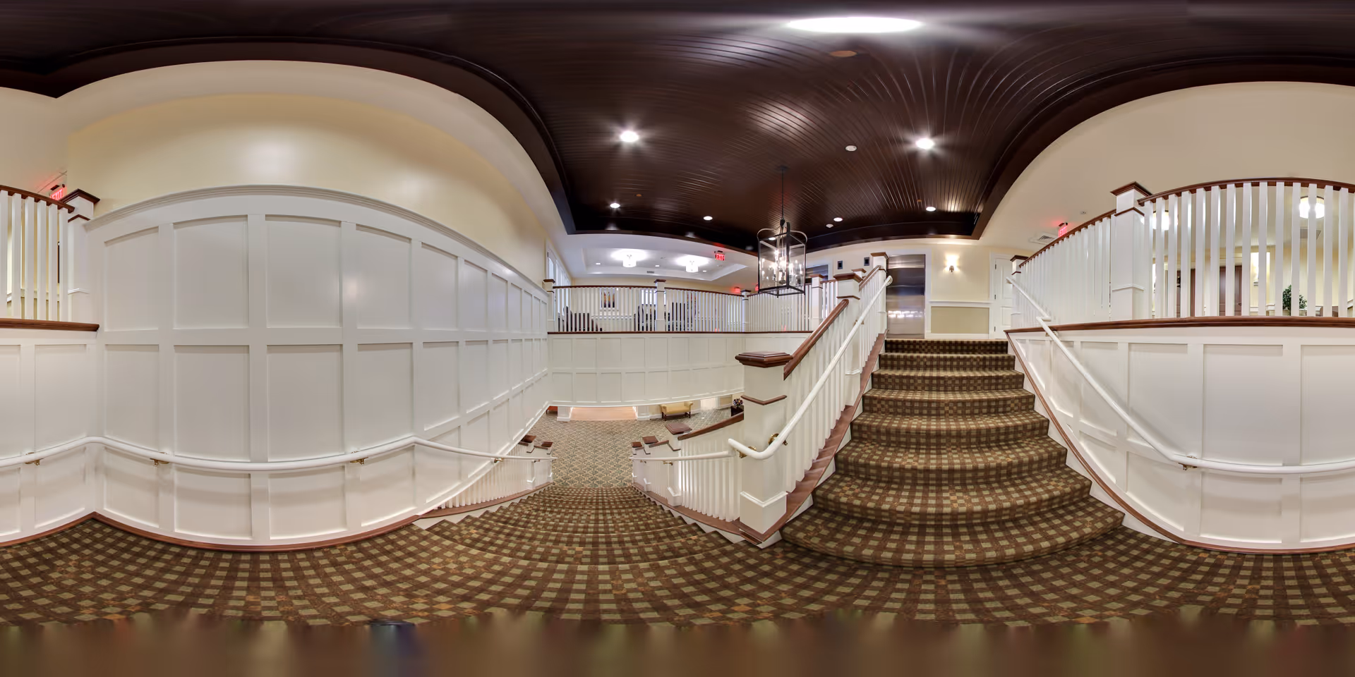 Interior view of a senior living facility featuring a carpeted staircase with patterned carpeting leading up to a landing. The walls are adorned with white wainscoting and the ceiling is dark wood with recessed lighting. Handrails are present on both sides of the stairs, and there is a chandelier hanging above the landing area. The space appears clean, well-lit, and welcoming.