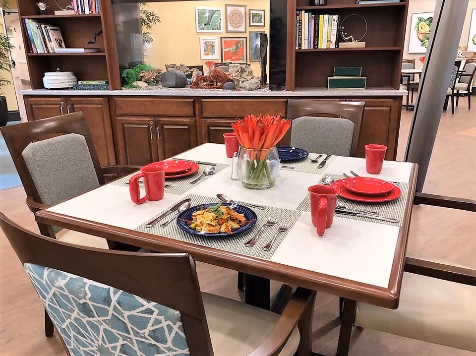 A dining table set for four in a senior living dining area with red plates and mugs, a vase of orange tulips, and chairs in front of a wooden cabinet and shelving.