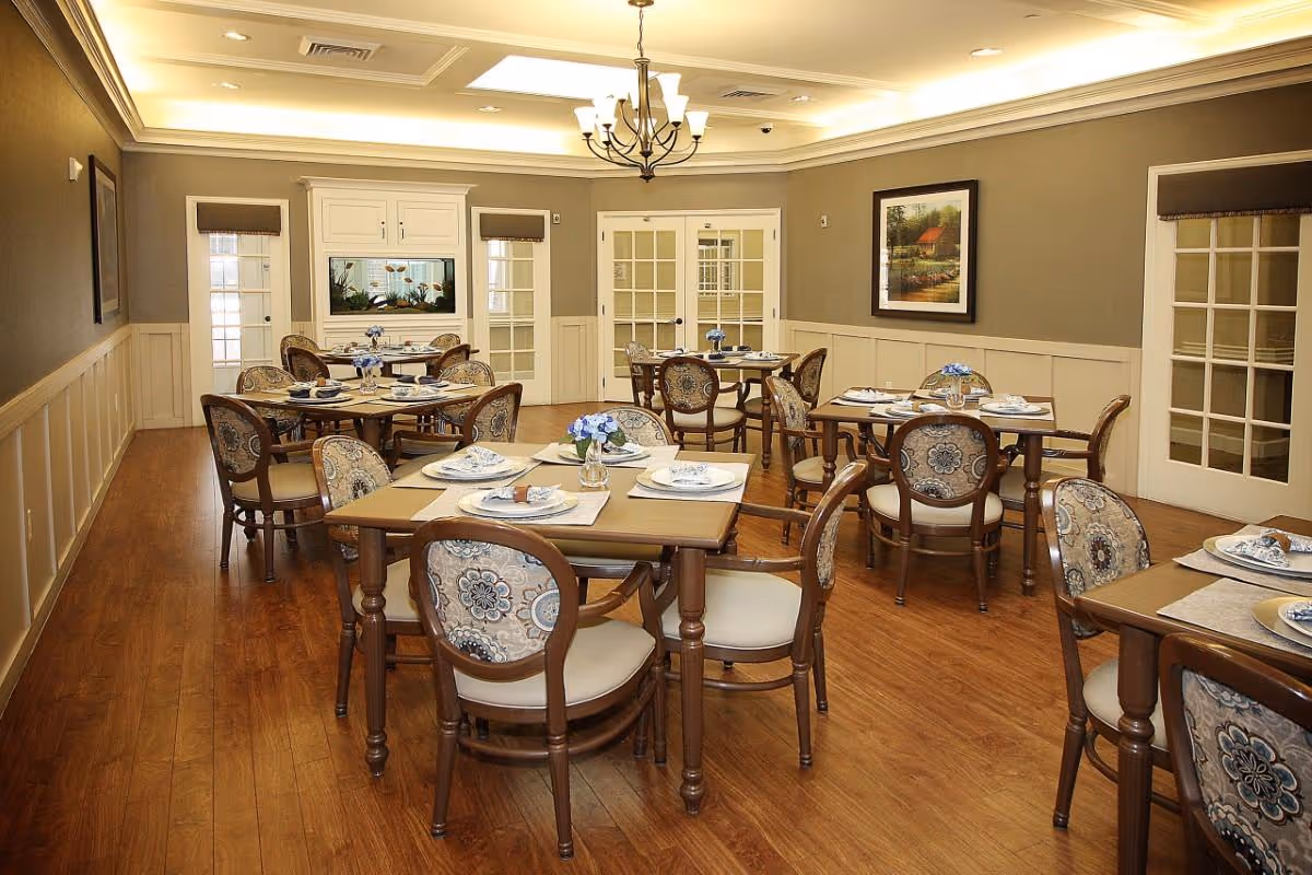 A dining room with several wooden tables and upholstered chairs arranged neatly. Each table is set with placemats, plates, napkins, and small flower vases. The room has wooden flooring, beige walls with white wainscoting, a chandelier hanging from the ceiling, and framed artwork on the walls. There are glass-paneled doors and windows letting in natural light.