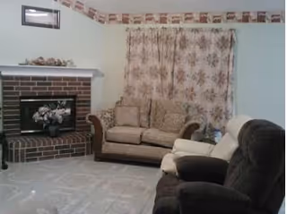 A cozy living room with a brick fireplace on the left, a floral arrangement on the mantel, a floral patterned curtain covering the window, a beige loveseat, and two recliner chairs, one beige and one dark brown.