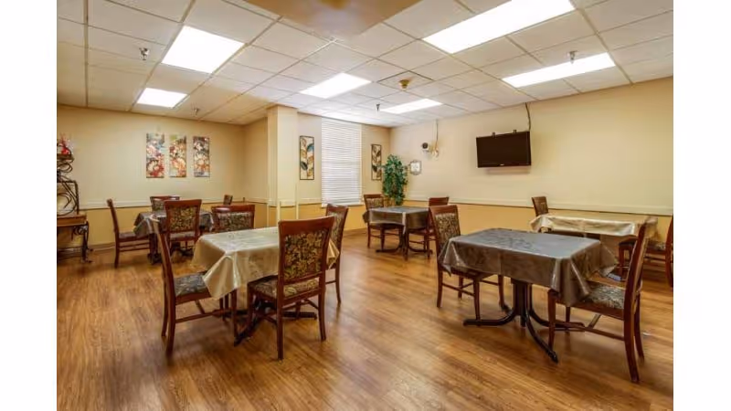 A dining room with several tables covered with tablecloths and surrounded by chairs. The room has wood flooring, beige walls, a window with blinds, decorative wall art, a mounted flat screen TV, and a potted plant in the corner.