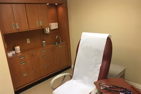 A medical examination room with a maroon examination chair covered with white paper, a wooden cabinet with multiple drawers and cupboards, a small sink, a soap dispenser, and a stethoscope placed on a nearby surface.