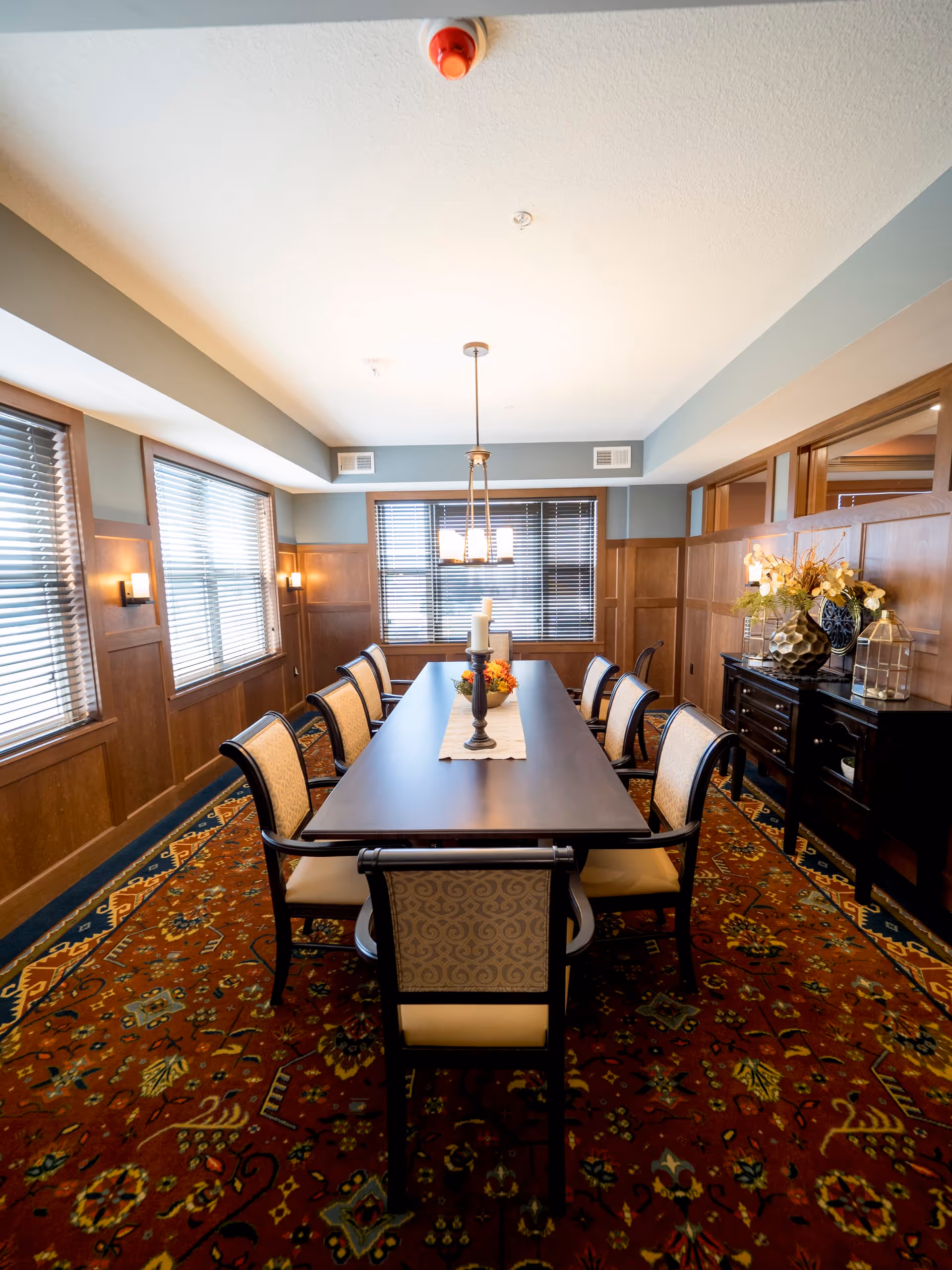 A formal dining room with a long rectangular wooden table surrounded by eight upholstered chairs. The room features wood-paneled walls, large windows with blinds, a patterned carpet, and a chandelier hanging above the table. A sideboard with decorative items and flowers is placed against one wall.