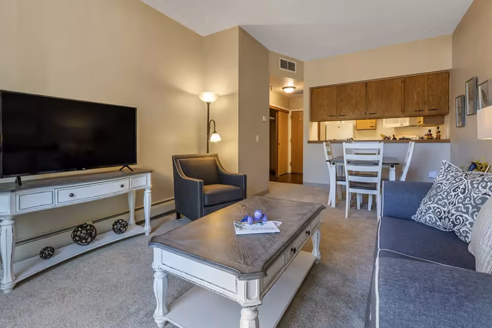 A cozy living room area with a gray sofa adorned with patterned pillows, a dark armchair, and a white wooden coffee table with decorative items on top. A flat-screen TV sits on a white console table against the wall. In the background, there is a dining area with a white table and chairs, and a kitchen with wooden cabinets and a pass-through window.