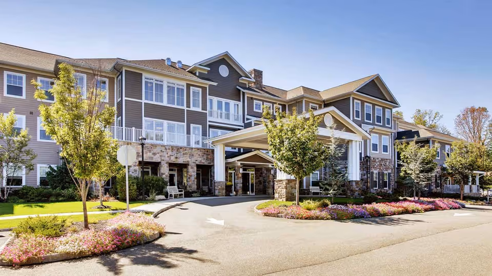 Exterior view of a large, multi-story senior living facility building with a covered entrance driveway. The building features a combination of stone and siding with numerous windows. There are landscaped flower beds and small trees along the driveway under a clear blue sky.
