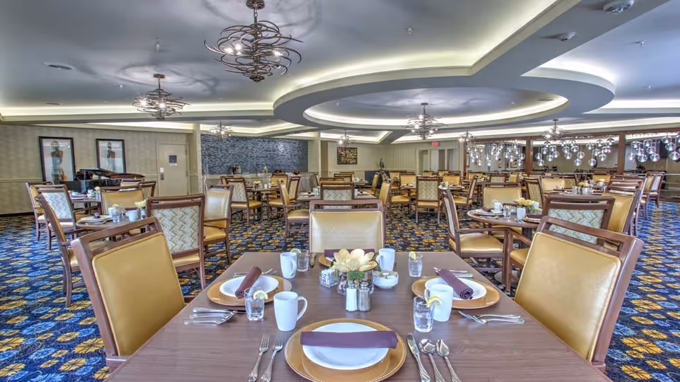 A spacious dining room with multiple tables set for four people each. The tables have plates, cups, glasses with lemon slices, silverware, and folded napkins. The room features patterned carpet, decorative ceiling lights, and framed artwork on the walls.
