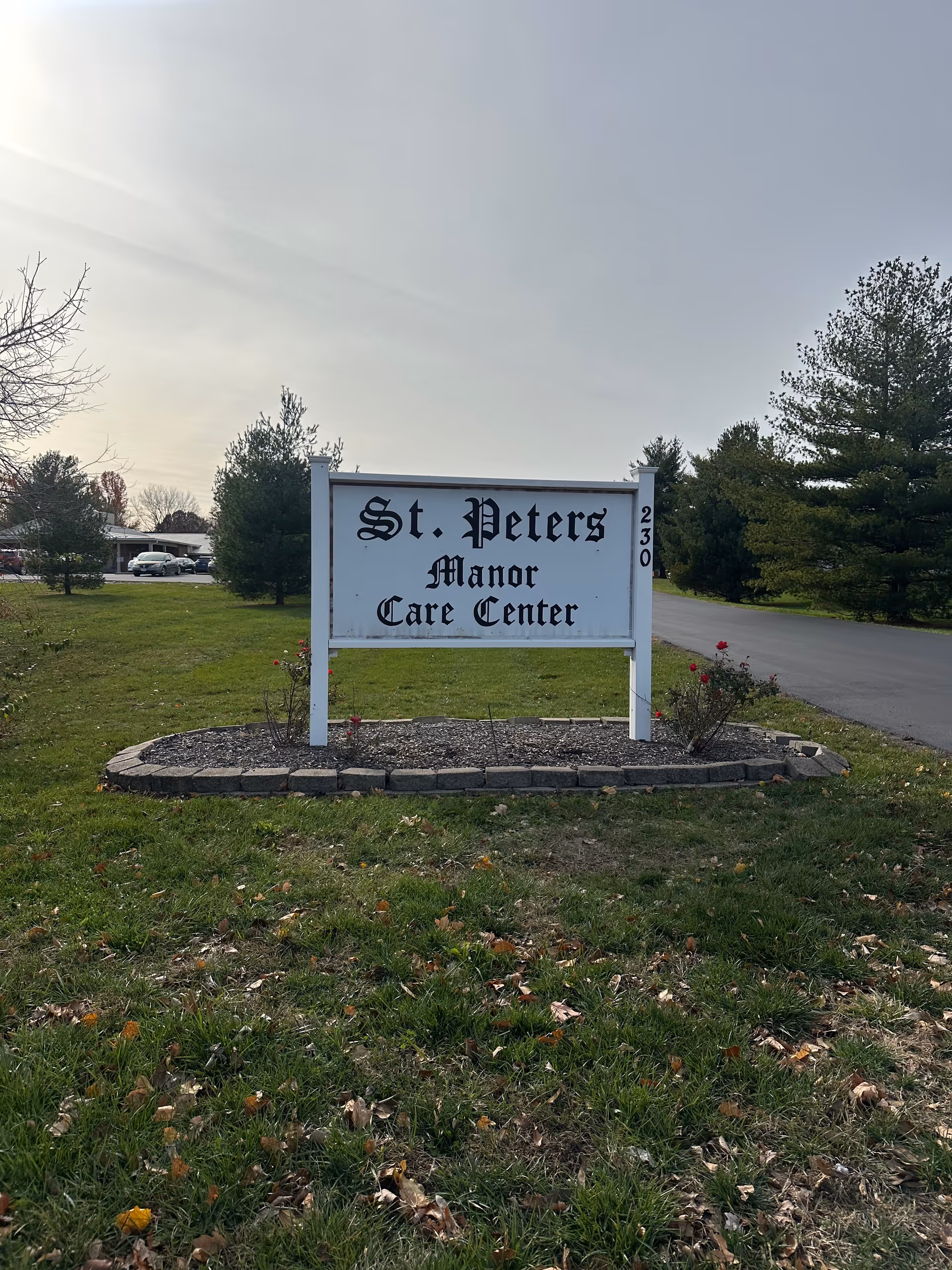 White roadside sign reading 'St. Peters Manor Care Center' on a grassy lawn with trees and a driveway in the background.