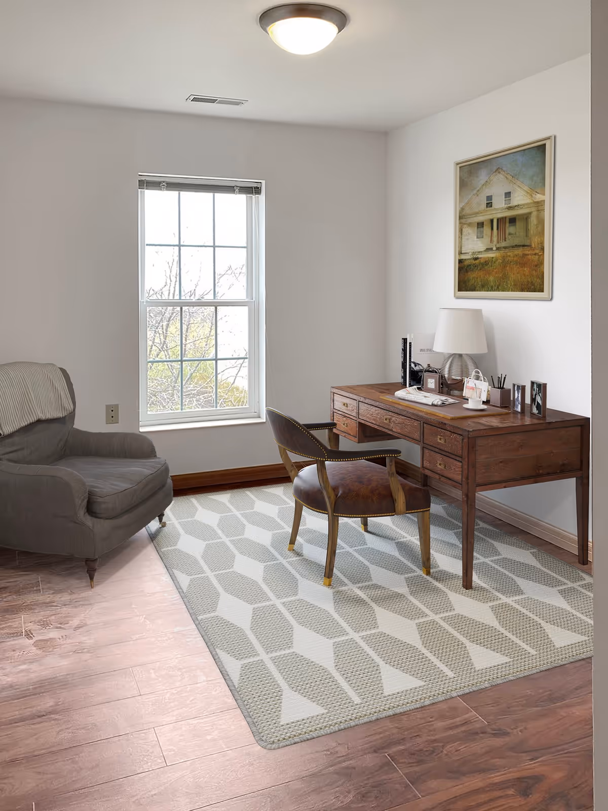A cozy room with a large window letting in natural light, featuring a wooden desk with multiple drawers, a leather chair, a table lamp, books, and framed photos on the desk. There is a patterned area rug on the wooden floor and a comfortable armchair with a blanket draped over the back. A framed picture of a house hangs on the wall above the desk.