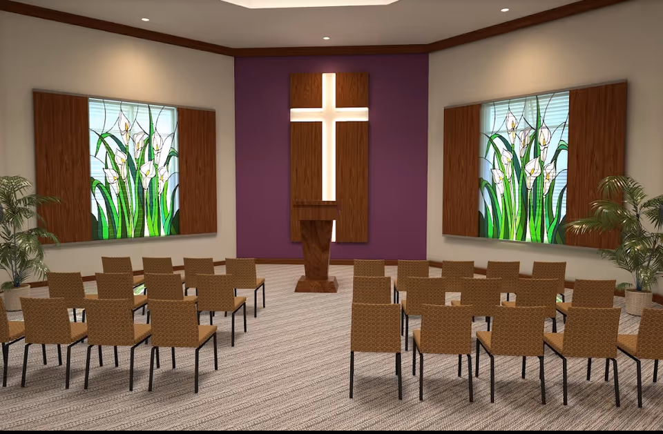 Chapel-style interior with rows of chairs facing a wooden lectern and illuminated cross, flanked by stained-glass windows and potted plants.