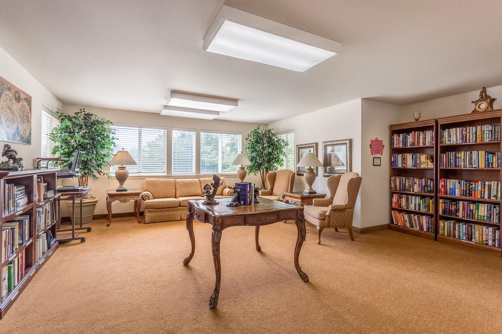 A cozy living room area with beige carpet and walls, featuring a wooden coffee table in the center with decorative items and books on it. There are two beige upholstered armchairs and a matching sofa along the walls. Large windows with blinds let in natural light. Two large potted plants and two table lamps on wooden side tables add to the decor. Bookshelves filled with books line the walls on the left and right sides of the room.