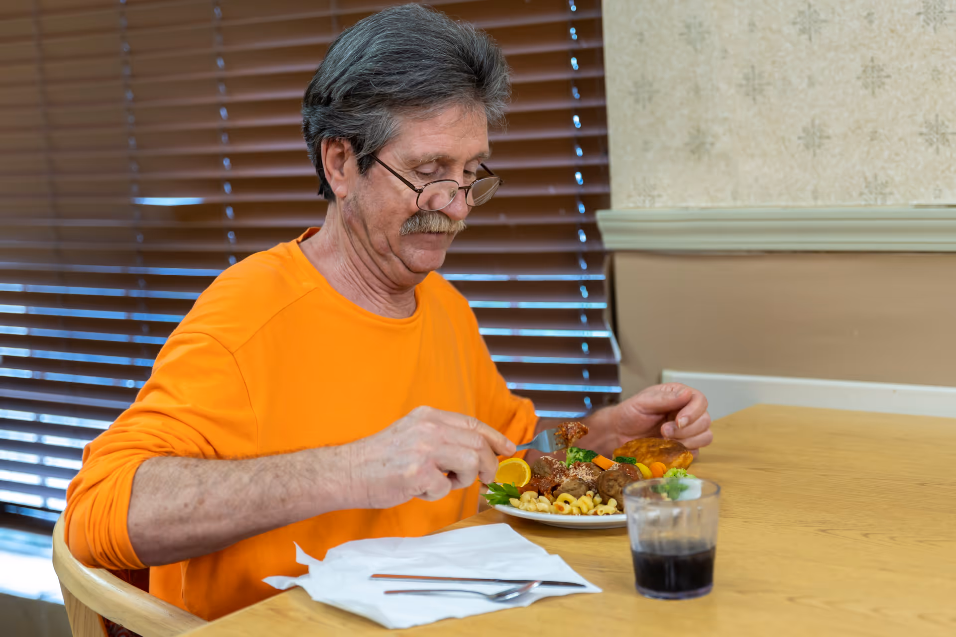 An elderly man wearing an orange shirt and glasses is sitting at a wooden table in a dining area, eating a meal that includes pasta, meatballs, vegetables, and a bread roll. There is a glass of dark beverage on the table, and the background shows closed wooden blinds and a beige wall.