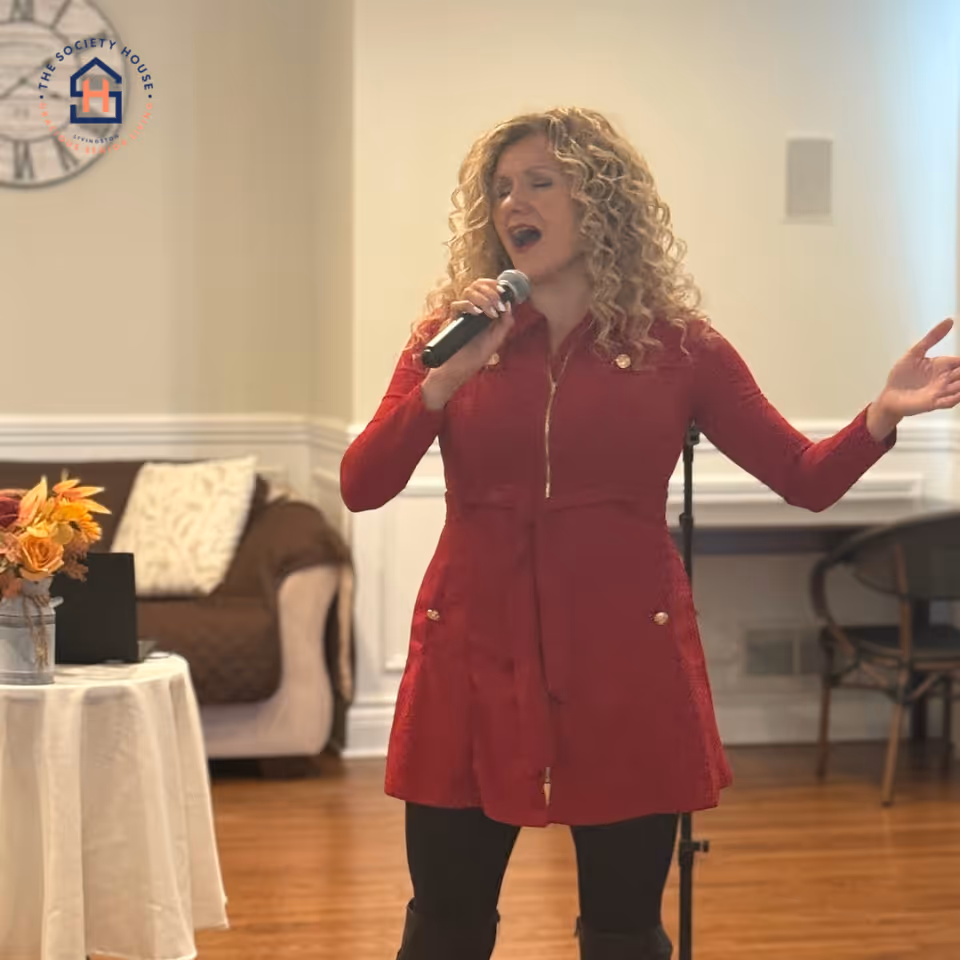 A woman with curly blonde hair wearing a red dress is singing into a microphone in a cozy indoor setting with wooden floors, a small table with a flower arrangement, and a couch with a pillow in the background.