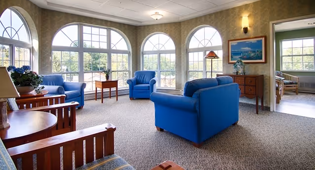 Sunlit common lounge with blue upholstered chairs, wooden side tables, and large arched windows.