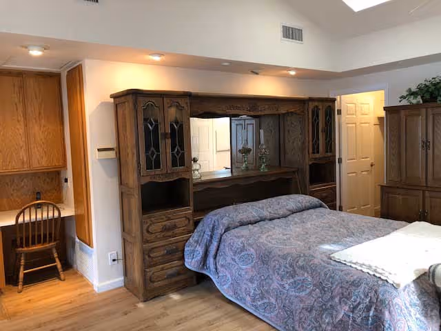 A bedroom with a wooden bed frame that includes built-in cabinets and shelves. The bed is covered with a patterned blue and purple bedspread. To the left, there is a small wooden desk with a chair and overhead cabinets. The room has light-colored walls and wooden flooring. A doorway is visible in the background, and there is a cabinet with plants on top to the right.