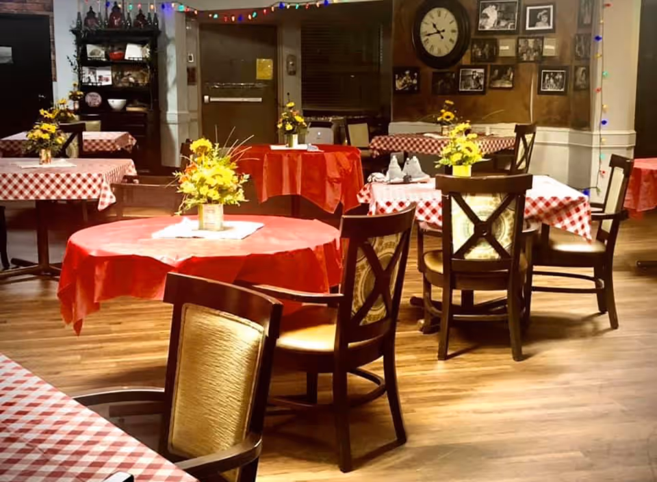 A cozy dining room with wooden floors and several tables covered with red and white checkered tablecloths and red tablecloths. Each table has a small vase with yellow flowers. Wooden chairs with cushioned seats surround the tables. The walls feature a large clock and several framed black and white photographs. Colorful string lights hang along the walls, adding a festive touch.