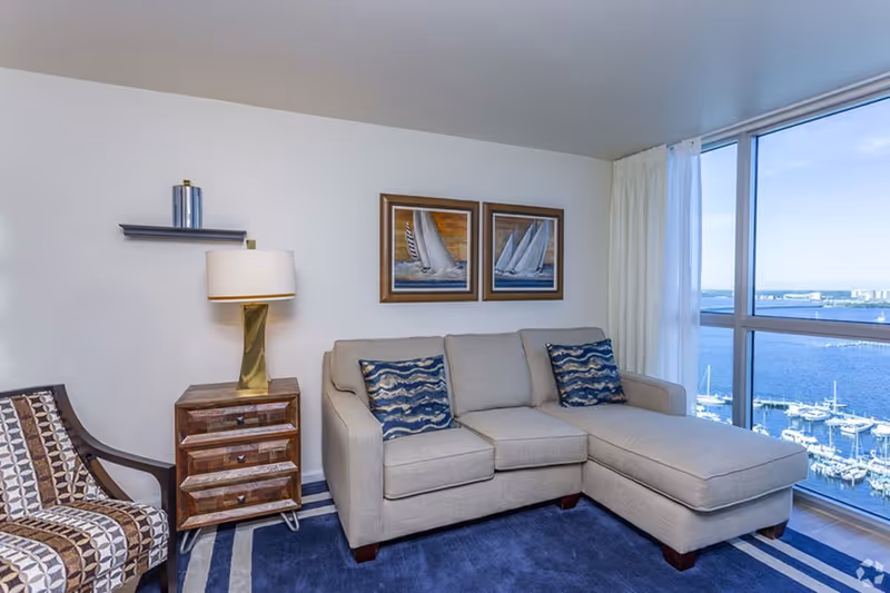 Bright living room with a beige sectional sofa, patterned chair, side table and large window overlooking a marina.