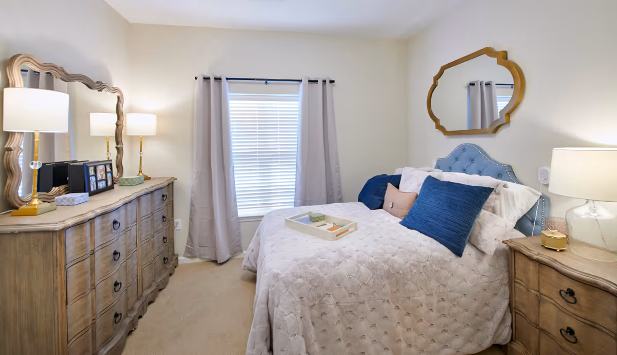 A cozy bedroom with a bed covered in a white textured comforter and blue accent pillows. There is a wooden dresser with a large decorative mirror and two lamps on the left side of the room. A window with closed blinds and light gray curtains is centered on the back wall. On the right side, there is a wooden nightstand with a lamp and a decorative mirror hanging above the bed's tufted blue headboard.