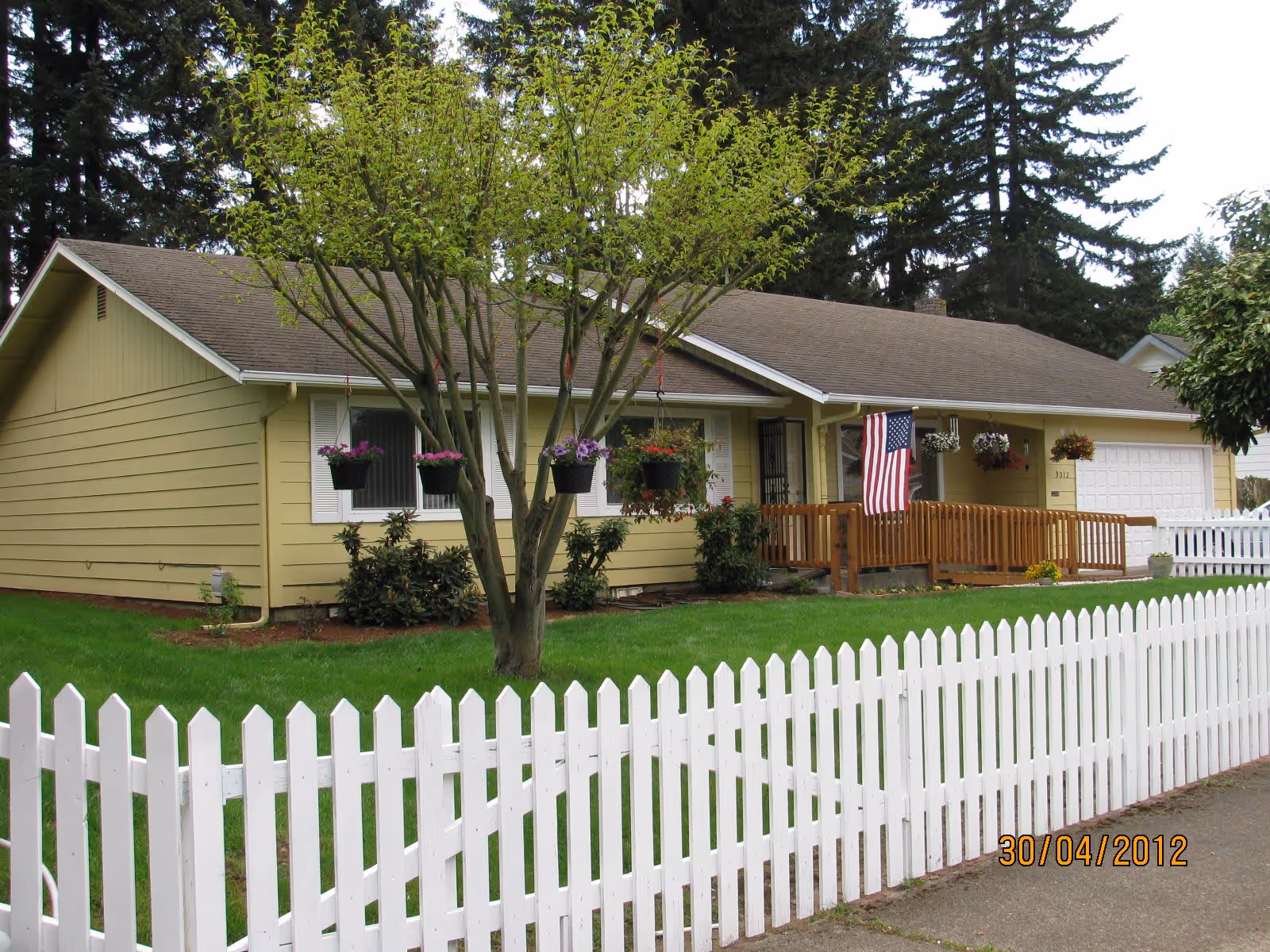 Single-story yellow house with a brown roof, a white picket fence in the foreground, a tree with hanging flower pots in the front yard, an American flag near the entrance, and a wooden ramp leading to the front door.