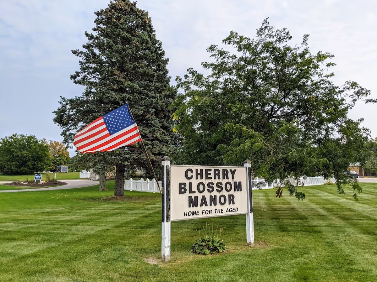 Outdoor view of a green lawn with a sign that reads 'CHERRY BLOSSOM MANOR HOME FOR THE AGED' next to an American flag on a pole. There are trees and a driveway in the background under a cloudy sky.