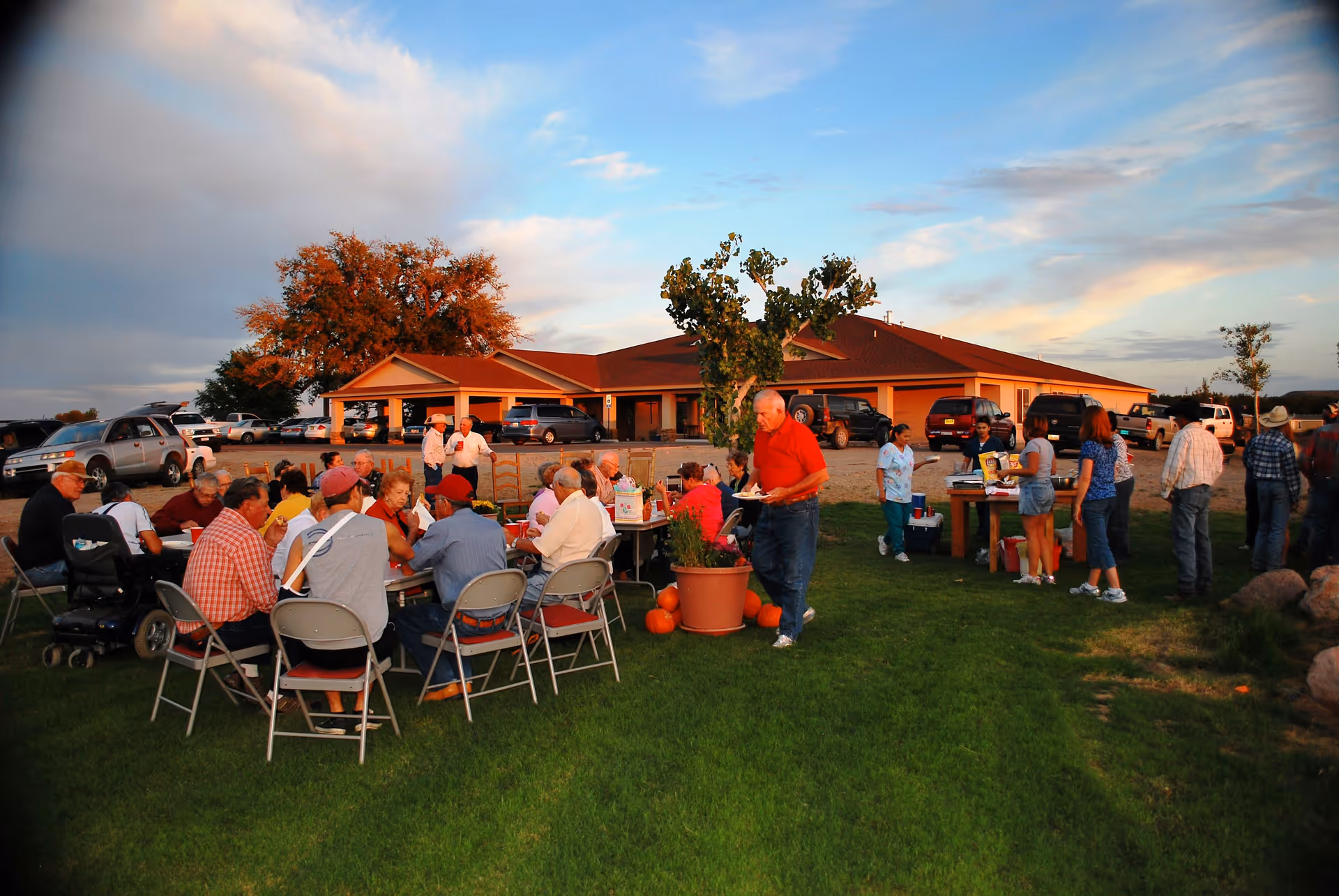 A group of elderly people and some younger individuals gathered outdoors on a grassy area near a building with a red roof. Several people are seated around tables eating and socializing, while others stand in line near a table with food and drinks. Cars are parked in the background under a partly cloudy sky during what appears to be late afternoon or early evening.