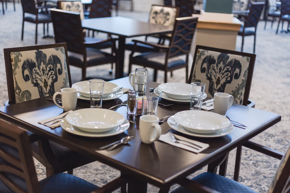 A dining table set for four with white plates, bowls, glasses, coffee mugs, and silverware in a senior living facility dining room. The chairs have patterned upholstery and there are more tables and chairs visible in the background.