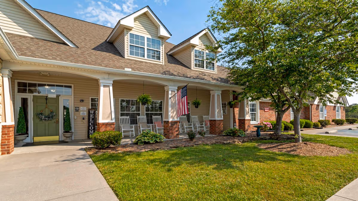 Front exterior view of Brookdale Kingsport facility showing a welcoming porch with rocking chairs, hanging plants, an American flag, and a green lawn with trees and shrubs under a blue sky.