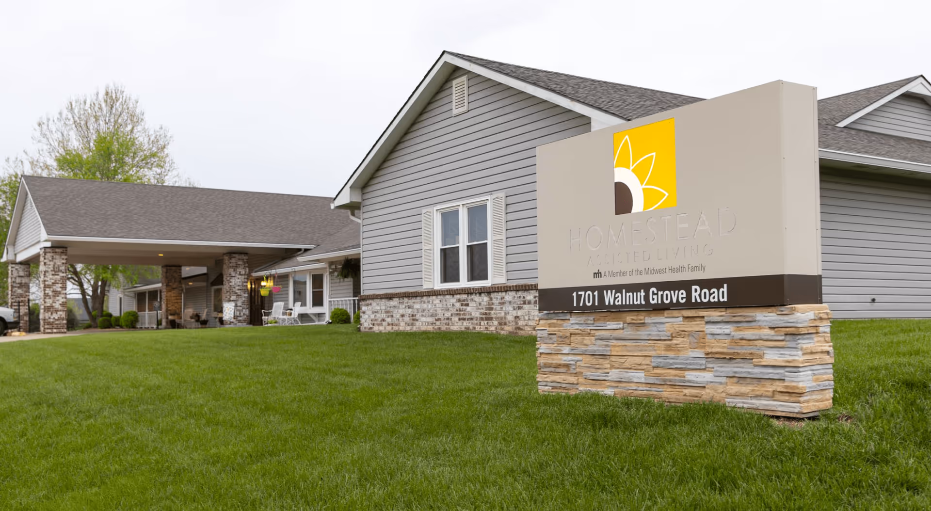 Exterior view of Homestead Assisted Living of Derby showing a single-story building with gray siding and a covered entrance supported by stone pillars. In the foreground, there is a large sign with the facility's name, logo, and address 1701 Walnut Grove Road, set on a stone base with green grass surrounding it.