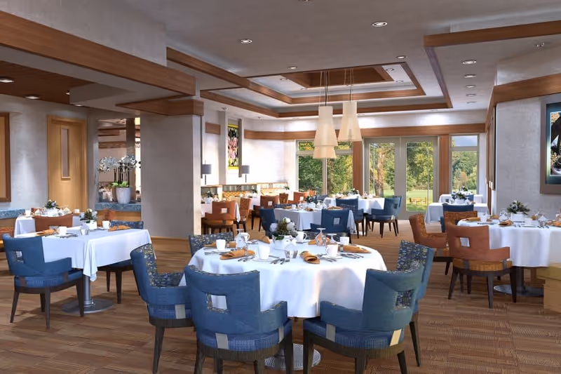 A spacious and well-lit dining room with round tables covered in white tablecloths, set with plates, glasses, and napkins. The room features a mix of blue and orange cushioned chairs, wooden flooring, large windows offering a view of greenery outside, and modern ceiling lights.