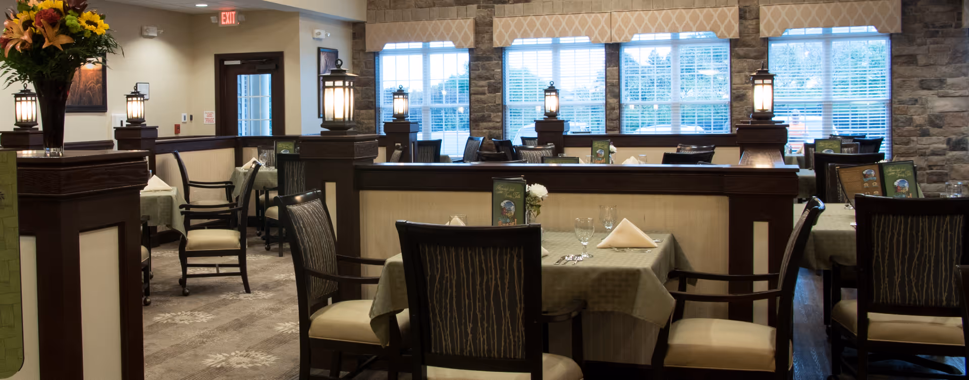 A dining room in a senior living facility with tables covered in green tablecloths, set with folded napkins, glasses, and silverware. The room features wooden chairs with cushioned seats and backs, decorative lantern-style lights on wooden dividers, large windows with beige valances, and a stone accent wall.