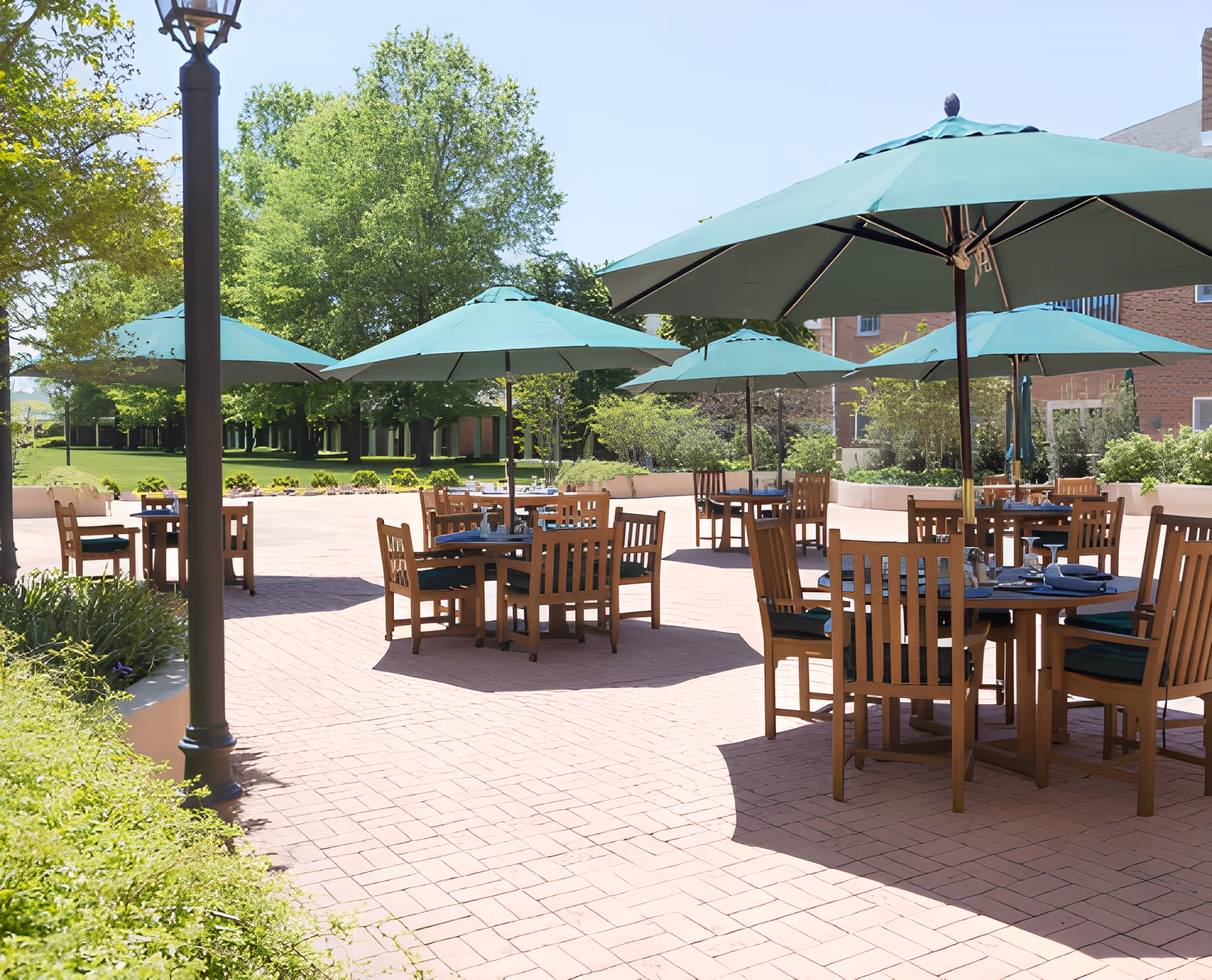 Outdoor patio area with multiple wooden tables and chairs, each shaded by large green umbrellas. The patio is paved with brick and surrounded by greenery and trees, with a building visible in the background under a clear blue sky.