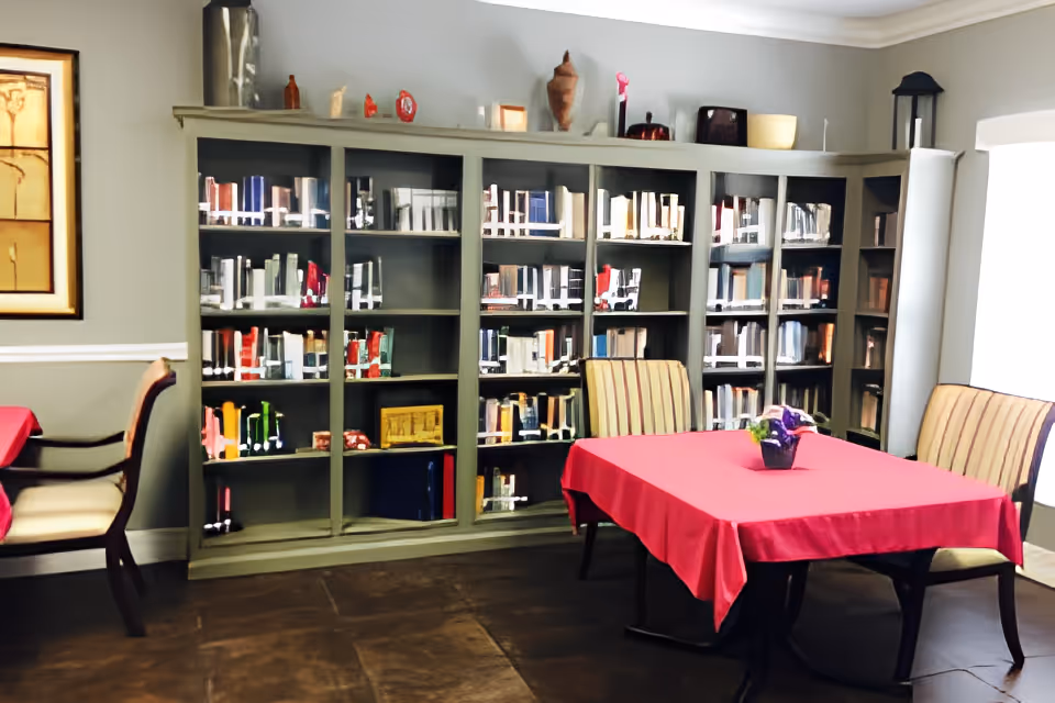 A cozy room with a large bookshelf filled with books along the wall, a table covered with a red tablecloth, and two striped upholstered chairs. The room has neutral-colored walls, a framed artwork on the left wall, and a window with white curtains on the right side.