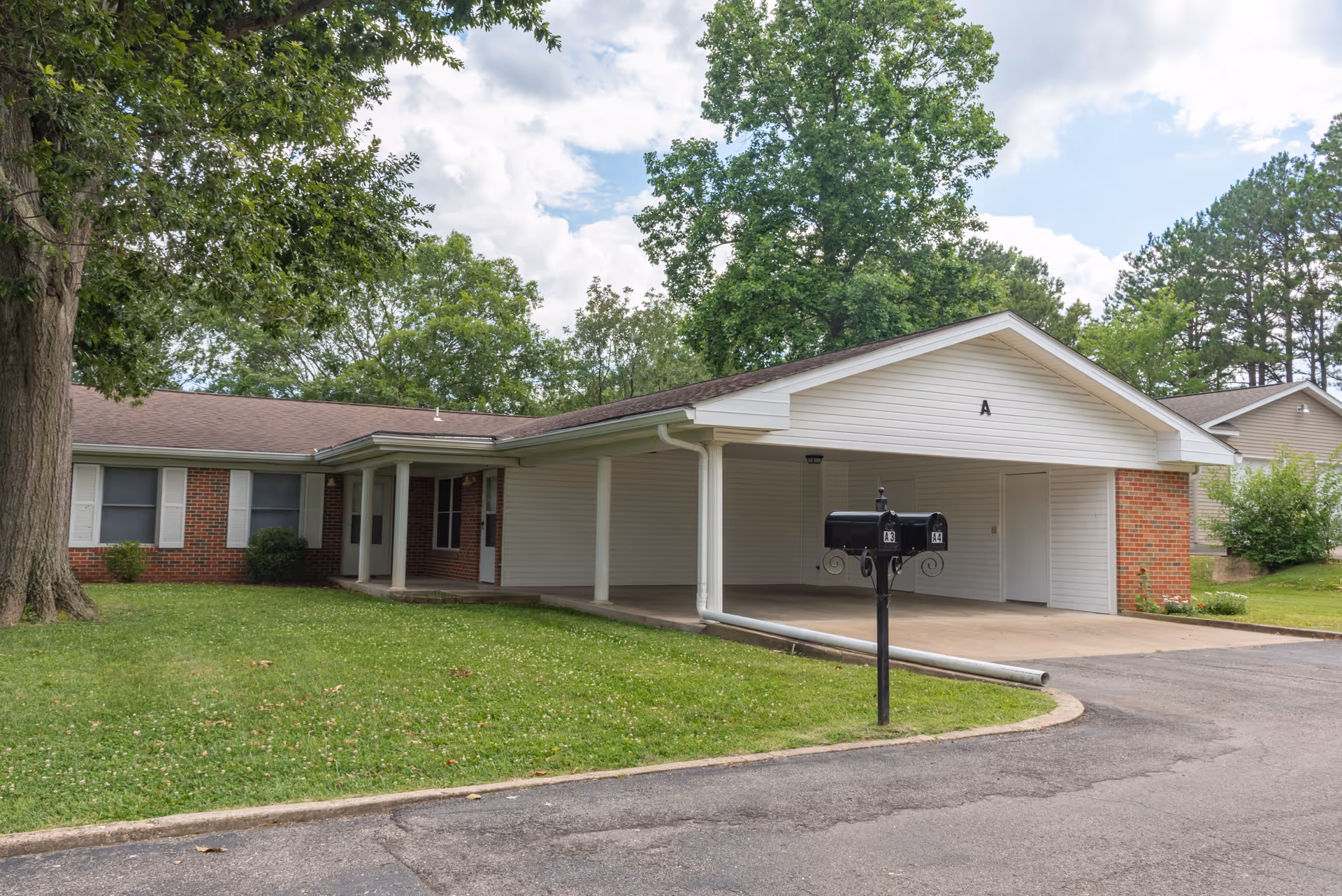 Single-story residential building with a covered carport labeled 'A', surrounded by green grass and trees under a partly cloudy sky. There are two black mailboxes on a post near the driveway.