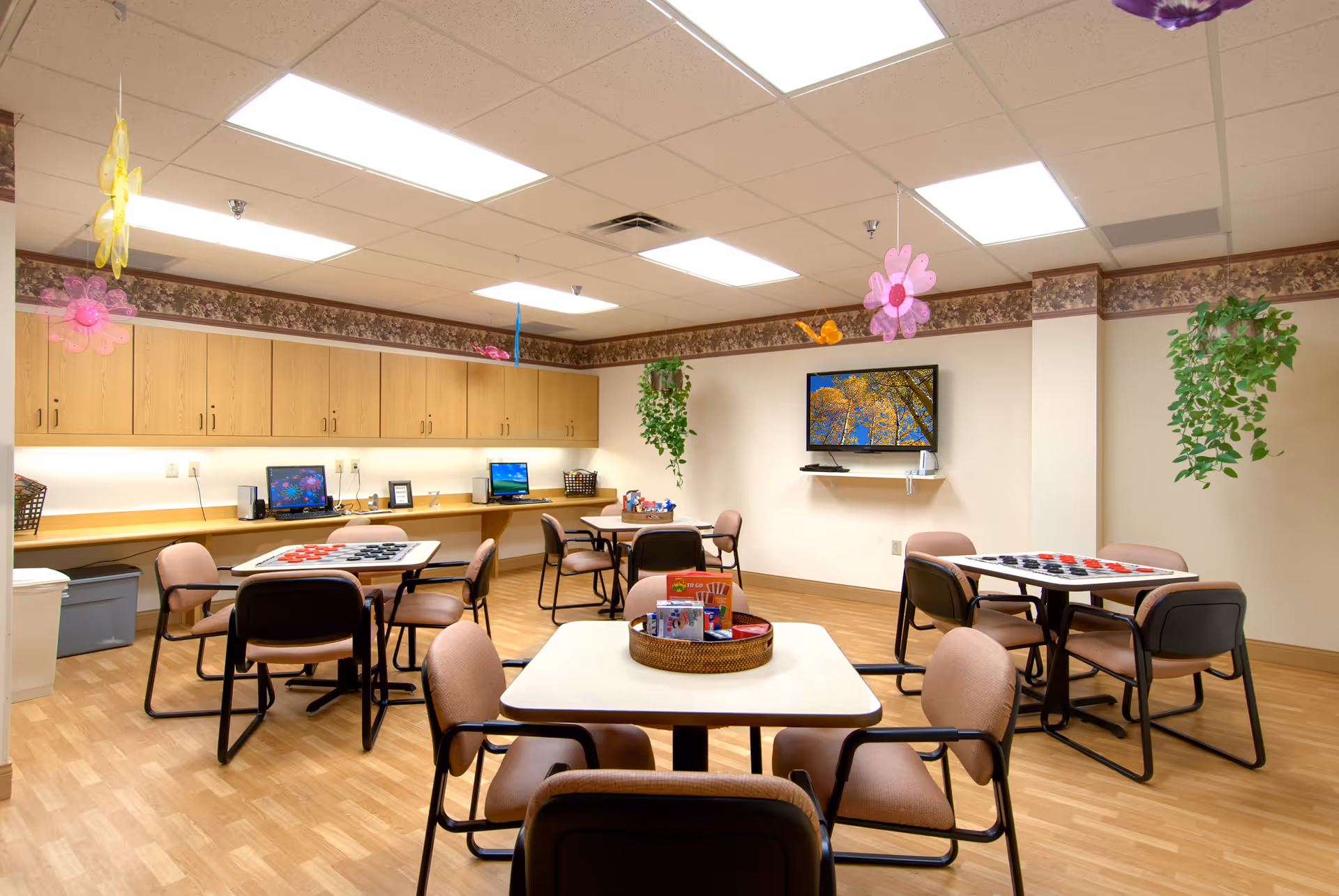 A bright and clean activity room with several tables and chairs arranged for group activities. The tables have board games like checkers set up. There are computers along the back wall under wooden cabinets, and a flat-screen TV mounted on the wall displaying an image of trees with autumn leaves. The room is decorated with hanging colorful flower decorations and green plants.