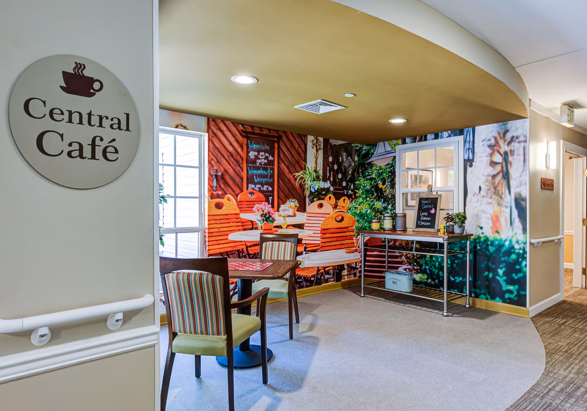 Indoor seating area labeled Central Café with a small table and two chairs in front of a large wall mural depicting an outdoor café scene with orange chairs and greenery. A metal cart with plants and a small chalkboard sign is placed against the mural wall. The area has a beige and light yellow color scheme with overhead lighting.