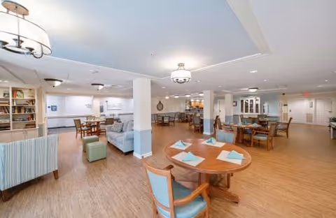 Spacious open common area with dining tables, chairs, lounge seating and bookshelves under soft overhead lighting.