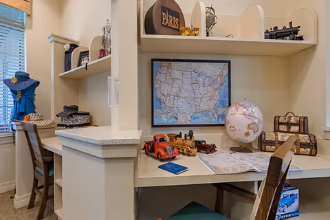 A cozy interior workspace with two desks separated by a partial wall. The right desk features a globe, a map, toy vehicles including a red truck and a train, a framed map of the United States on the wall, and decorative storage boxes. The left desk has a chair, a hat stand with hats, and shelves with various decorative items. Both desks have wooden chairs with green cushions.