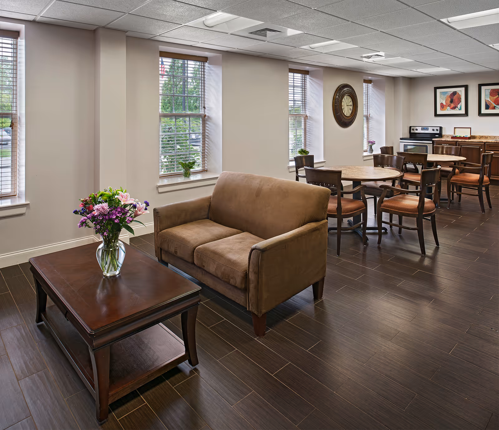 A cozy common area in a senior living facility featuring a brown loveseat, a wooden coffee table with a vase of colorful flowers, round tables with chairs, a wall clock, and a kitchen area with an oven and cabinets. The room has large windows with blinds and framed artwork on the wall.