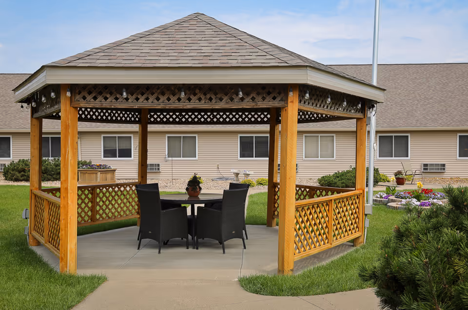 A wooden gazebo with a shingled roof situated on a concrete pad in a grassy outdoor area. Inside the gazebo, there is a round table with four black wicker chairs. In the background, there is a beige building with several windows and some landscaping including bushes and flower beds.