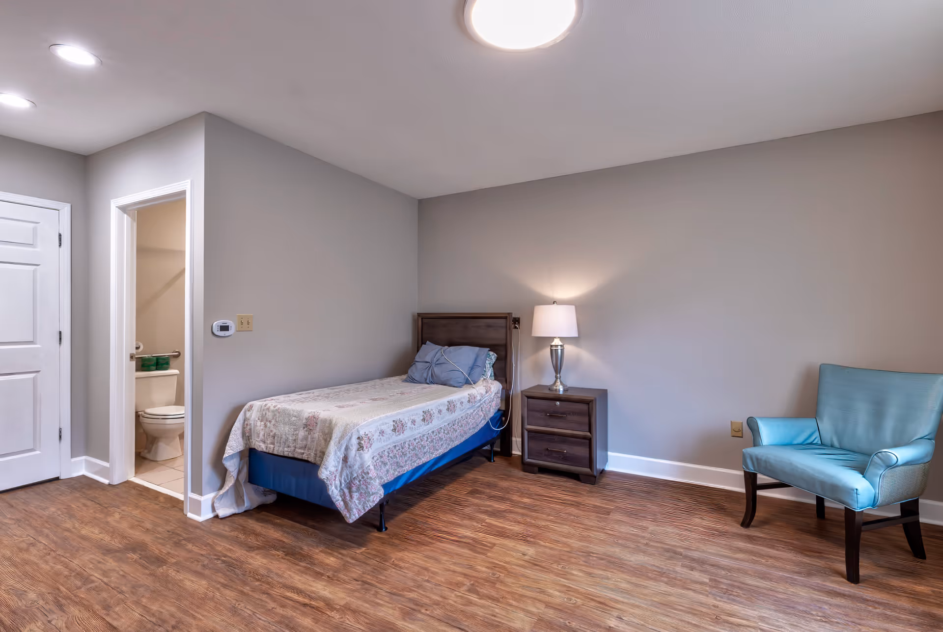 A simple bedroom with a single bed covered with a floral quilt, a wooden nightstand with a lamp, a light blue upholstered chair, and an open door leading to a bathroom with a visible toilet.