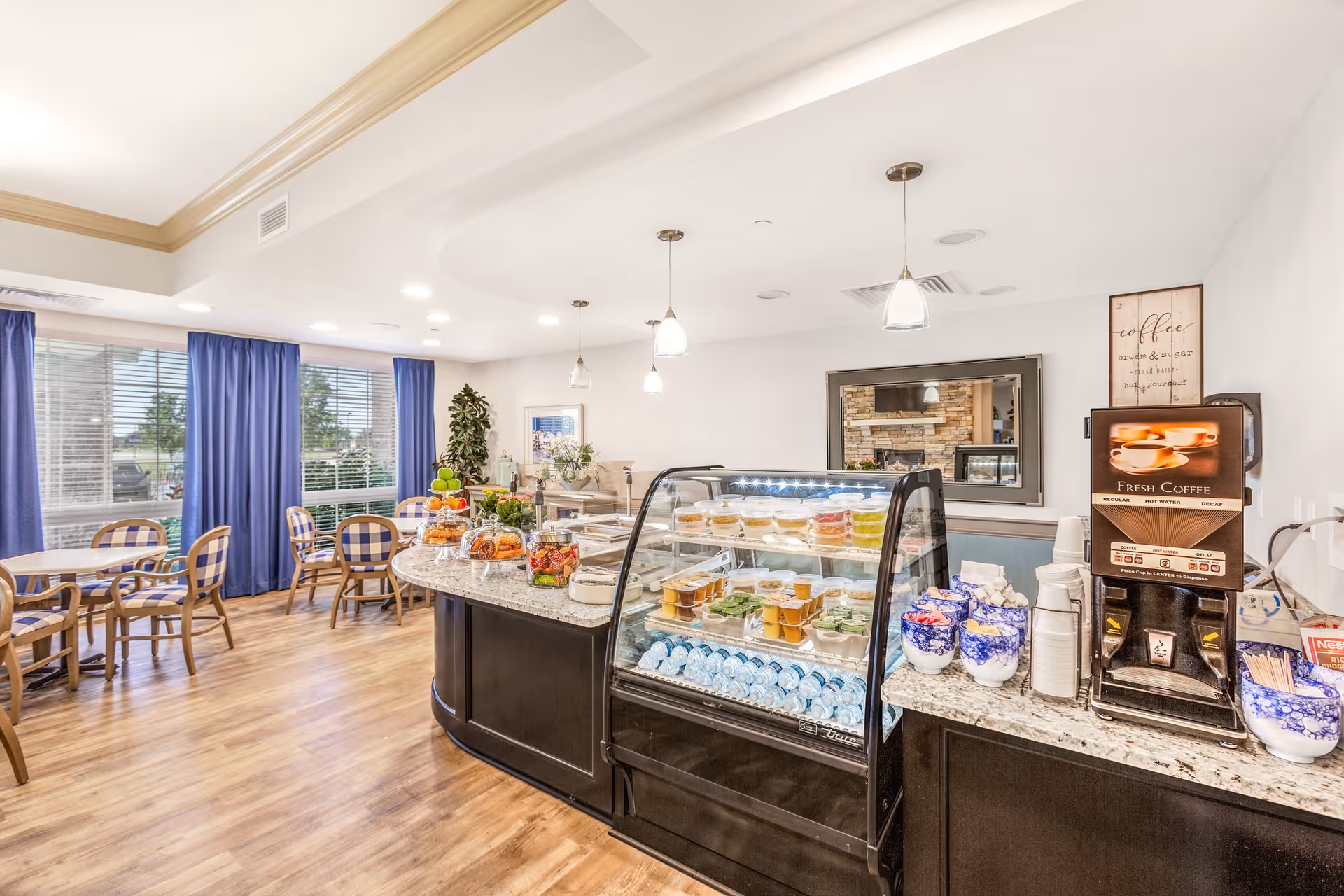 Bright dining area with wooden floors and several tables and chairs near large windows with blue curtains. A curved counter displays various food items in a glass case, with a coffee machine and cups on the side. The room is well-lit with ceiling lights and pendant lamps.