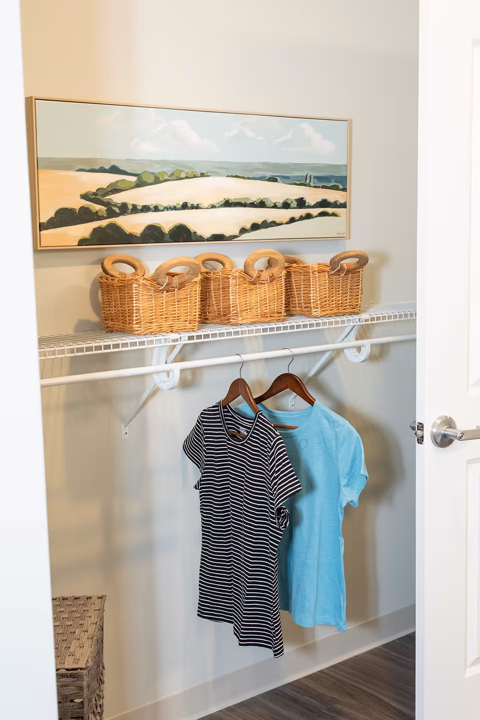 An open closet with a white wire shelf and hanging rod. On the shelf are four wicker baskets with wooden handles. Two shirts, one black and white striped and one light blue, hang on wooden hangers from the rod. Above the shelf is a framed landscape painting depicting fields and trees under a cloudy sky. The closet door is partially open, and the floor is wood.