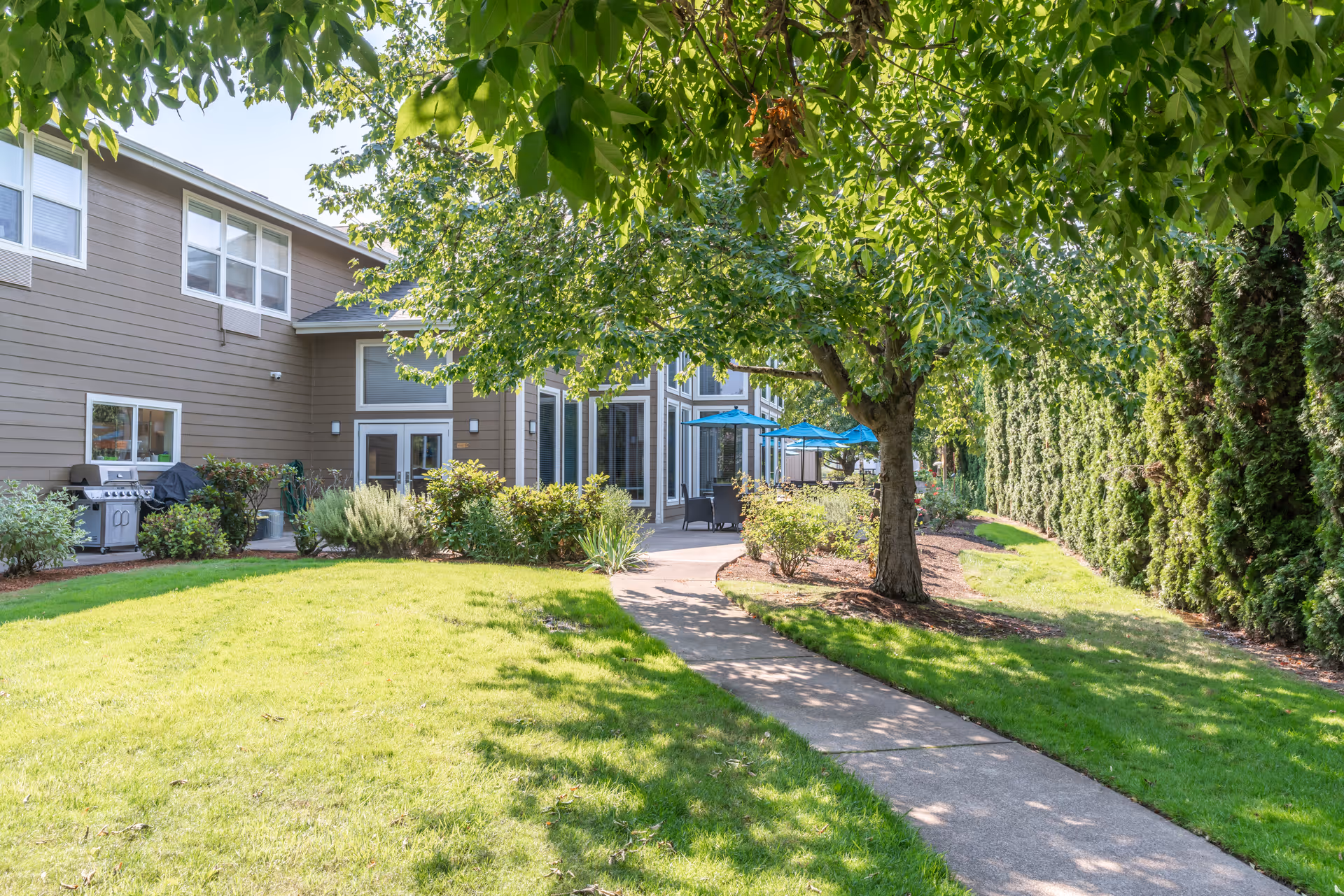 A paved walkway winds through a sunny grassy courtyard with trees, patio seating and blue umbrellas beside a two-story senior living building.