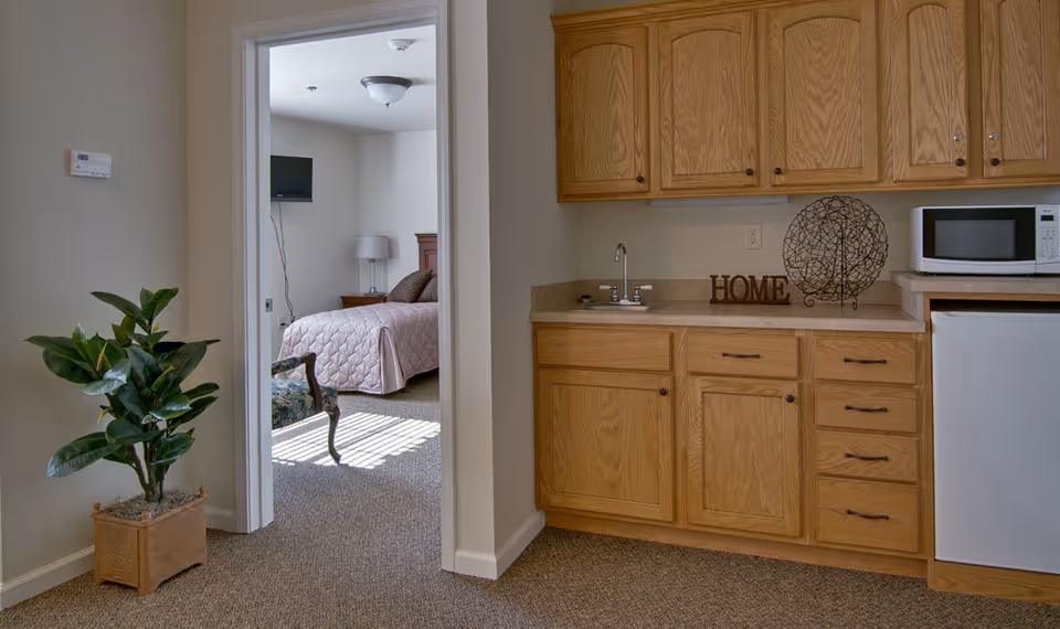 Small kitchenette with oak cabinets, sink and microwave, a doorway leading to a furnished bedroom, and a potted plant.