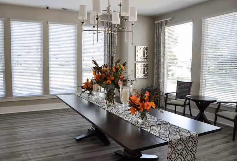 Sunlit communal dining room with a long dark wood table decorated with floral centerpieces and a chandelier.