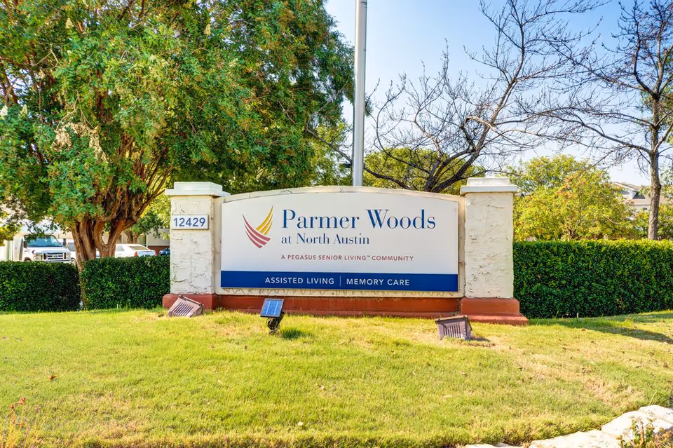 Outdoor view of a large sign for Parmer Woods at North Austin, a Pegasus Senior Living community offering assisted living and memory care, surrounded by green grass, trees, and bushes under a clear blue sky.