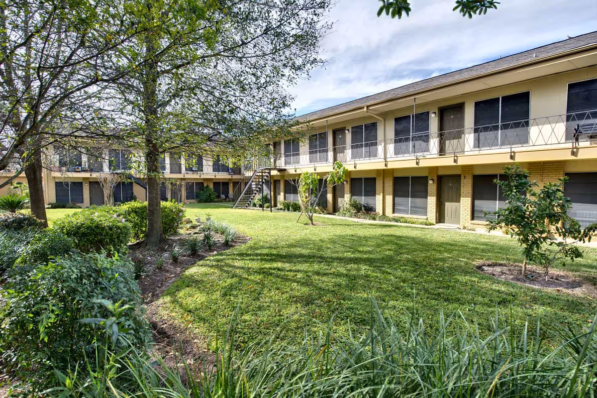 A two-story retirement community building surrounding a well-maintained grassy courtyard with trees, shrubs, and small plants. The building has multiple doors and windows with black screens, and an external staircase leads to the upper floor. The sky is partly cloudy.