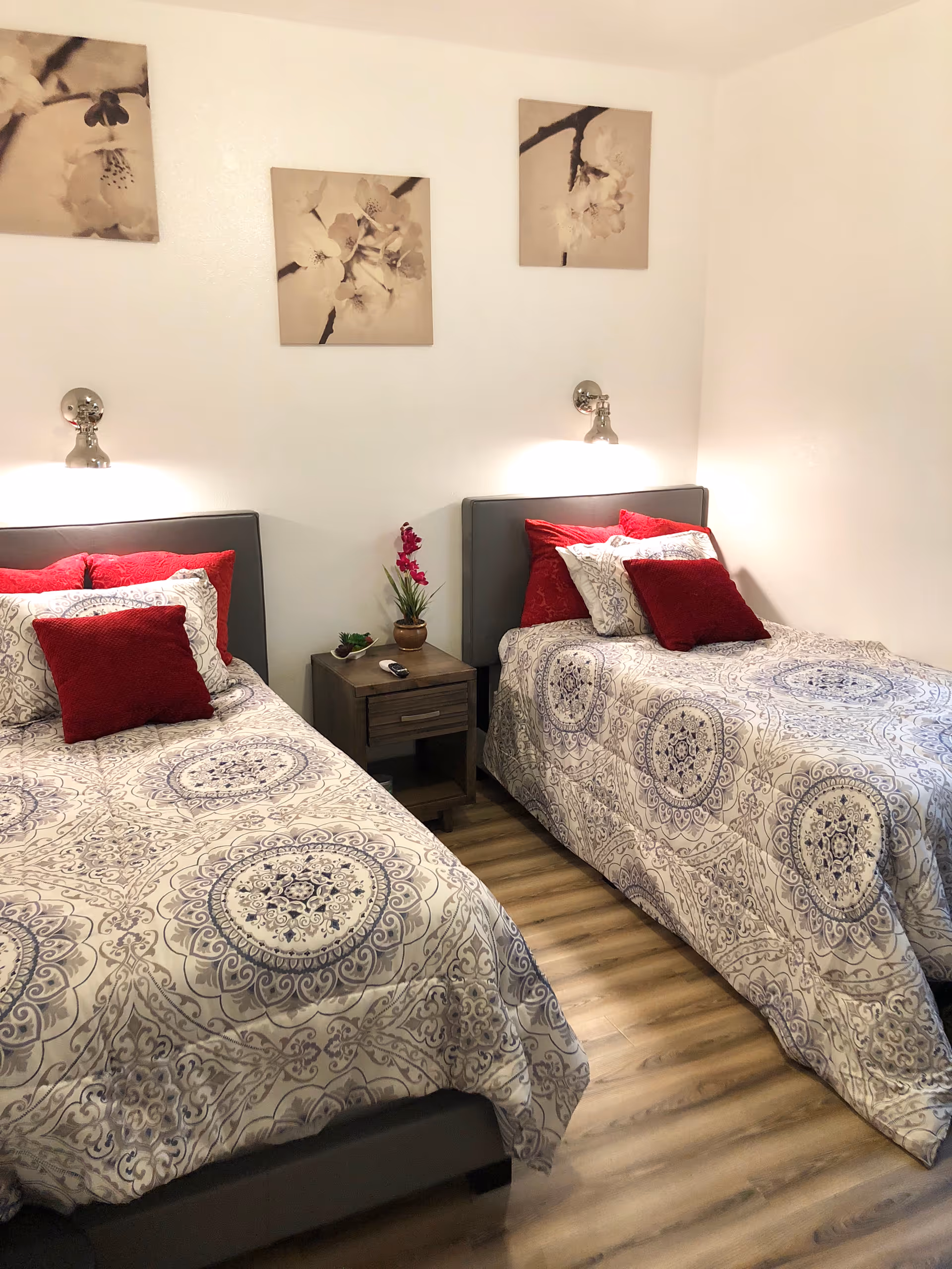 Two twin beds with patterned bedspreads and red accent pillows flank a small nightstand under floral wall art in a clean, well-lit bedroom.