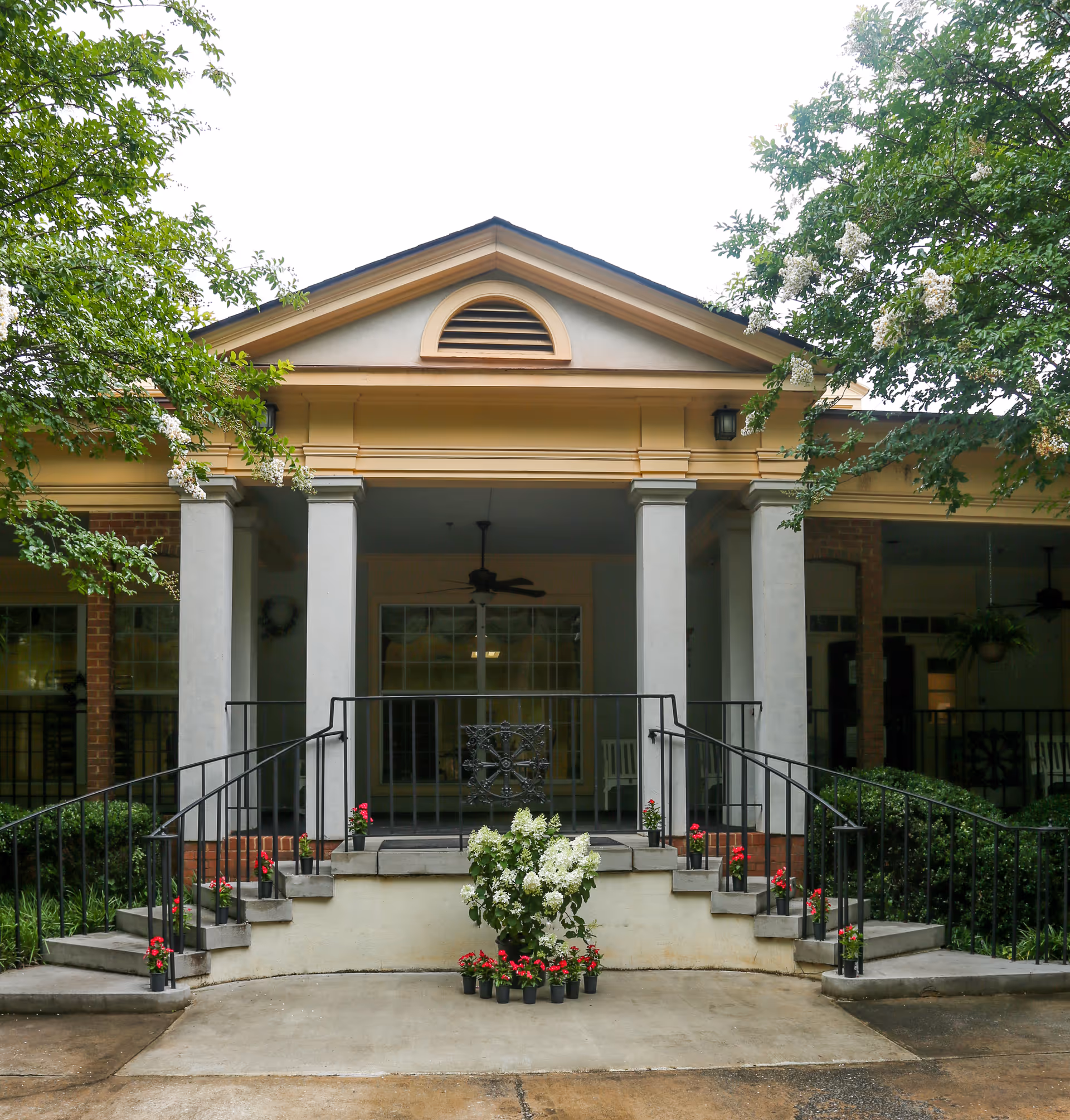Front entrance of a building with a small staircase on both sides leading up to a covered porch area. The porch has four columns and a ceiling fan, with potted flowers arranged along the stairs and at the base of the steps. Trees with green leaves and white flowers frame the entrance.